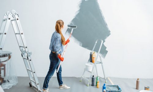 Female house painter with roller paints wall. Home repair, laughing woman doing appartment renovation, room decoration renovating
