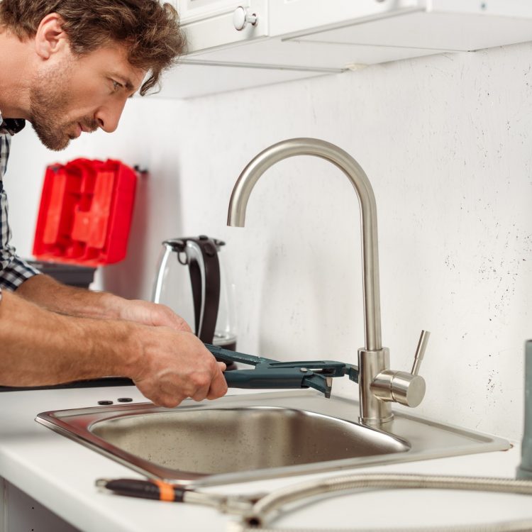 Side view of handsome repairman using pipe wrench while fixing kitchen faucet